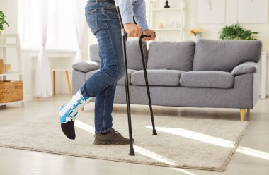 Evdeki oturma odasında koltuk değnekleriyle duran sakat ve kırık bacaklı bir gencin kesilmiş fotoğrafı. Bacağı alçılı özürlü adam. Rehabilitasyon ve iyileşme kavramı.