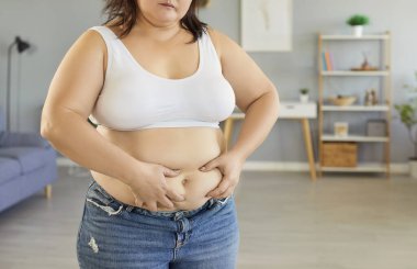 Şişman, şişman, şişman, günlük kıyafetler giyen genç bir kadının kesilip biçilmiş portresi. Evdeki oturma odasında, ne yazık ki koca karnına dokunuyor. Zayıflama, diyet, yazım kaybı ve obezite konsepti.