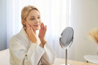 Kozmetik uygulayan güzel bir kadın, krem ürünüyle yüz yüze, aynaya bakıyor, beyaz bornoz giyiyor, cilt rutinini evde yapıyor, temizlik için kişisel bakım yapıyor, doğal yüz nemlendirici.