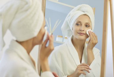 30 'lu yaşlardaki beyaz bornoz ve havlu giymiş genç bir kadın aynanın önünde duruyor, elinde pamuk tutarak yüzüne cilt bakım ürünleri uyguluyor. Kişisel bakım, kişisel bakım konsepti