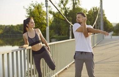 Güneşli bir günde göl kenarında kollarını ve bacaklarını birbirine uzatan zinde genç bir çift. Gülümseyen adam ve kadın spor kıyafetleriyle koşuya çıkmadan önce kaslarını ısıtıyor, aktif sağlıklı yaşam tarzının tadını çıkarıyor..