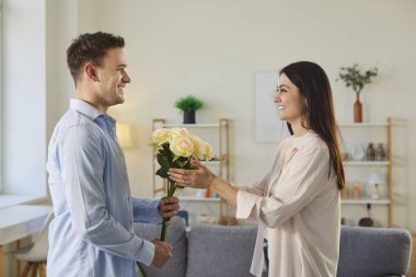 Genç, neşeli bir adam bir buket çiçekle tatil mutlu sevgilisini tebrik ediyor. Evdeki oturma odasında birbirine aşık, gülümseyen bir çift. Sevgililer günü ve ilişkiler konsepti.