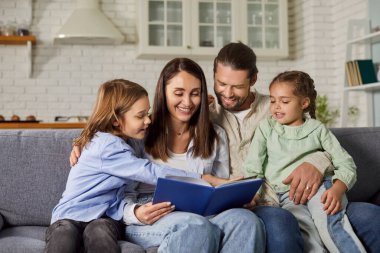 Kitap okuyan neşeli bir aile. Mutlu ebeveynler ve çocuklar ilginç bir hikayenin tadını çıkarıyorlar. Gülümseyen anne, baba ve çocuklar evlerindeki ya da evlerindeki kanepede oturup birlikte kitap okuyorlar.