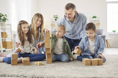 Oturma odasındaki yumuşak halıda oturan ve ahşap bloklarla kule inşa eden mutlu bir aile. Gülümseyen ebeveynler çocuklarını yaratıcı oynamaları için cesaretlendiriyor, evde kaliteli zaman geçirmekten ve takım çalışmasından keyif alıyorlar..