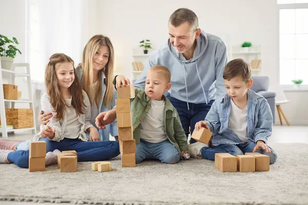 Oturma odasındaki yumuşak halıda oturan ve ahşap bloklarla kule inşa eden mutlu bir aile. Gülümseyen ebeveynler çocuklarını yaratıcı oynamaları için cesaretlendiriyor, evde kaliteli zaman geçirmekten ve takım çalışmasından keyif alıyorlar..