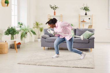 Neşeli Afro-Amerikalı kadın gündelik kıyafetlerini giyiyor ev işlerinde neşe buluyor, elektrikli süpürgeyi mikrofon olarak kullanıyor, eğleniyor, dans ediyor ve evde temizlik yaparken salonda şarkı söylüyor. Eğlenceli oda servisi..