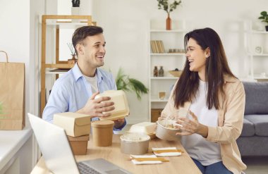 Yemek servisi yapan kafede hazır yemek ve paketleri açan mutlu evli çift. Genç bir adam ve bir kadın evde dizüstü bilgisayarlarıyla masada oturuyor ve plastik ve karton yiyecek konteynırlarını açıyorlar.