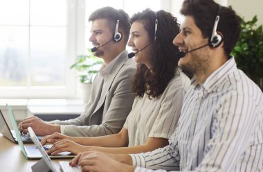 Üç gülümseyen genç çağrı merkezi operatörü sırada oturmuş müşteri destek ofisindeki dizüstü bilgisayarlarla uğraşıyorlar. Mesai saatlerinde müşterilerle iletişim kuran mutlu pazarlamacı takımı..