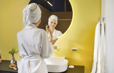 Genç bir kadının yuvarlak aynaya bakarken banyoda cilt kremi sürüşünü. Beyaz bornoz ve havlu giymiş, sabah rutininde nemlendirici kullanan gülümseyen bir kadın..