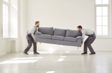 Üniformalı iki nakliyeci büyük kanepeyi kaldırıp boş odaya getiriyorlar. Müşteri için ev taşınması ya da teslimat hizmetleri sırasında mobilya taşımaya yardım eden profesyonel işçiler.