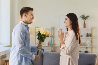 Genç, neşeli bir adam bir buket çiçekle tatil mutlu sevgilisini tebrik ediyor. Evdeki oturma odasında birbirine aşık, gülümseyen bir çift. Sevgililer günü ve ilişkiler konsepti.