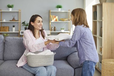 Evdeki oturma odasındaki kanepede oturan genç ve güzel bir kadın ve çocuk. Elinde bir sepet taze kumaş kıyafetle. Oğlum, neşeli annesine çamaşır yıkamasında yardım ediyor. Aile işleri ve ev işleri..