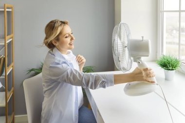 Elektrikli vantilatör kullanan bir kadın, havalandırmadan gelen temiz havanın tadını çıkarmak için serin hava kullanan bir kadın, ev aletleri, soğutma ekipmanları, kapalı havayı geliştiren bir kadın, sıcak hissetmeyen bir kadın.