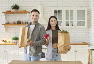 Mutfakta birlikte durup kameraya bakan, alışveriş poşetleri ve sebzelerle birlikte paketleri açan mutlu, genç bir çiftin portresi. Alışveriş, sağlıklı gıda ve beslenme konsepti.