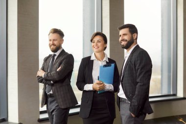 Ofiste yan yana duran üç iş adamı, yüzlerinde geniş bir gülümsemeyle gözlerini kaçırıyorlar. Siyah takım elbiseli kadın, iki neşeli, kendinden emin erkek meslektaşının yanında duran bir pano tutuyor..