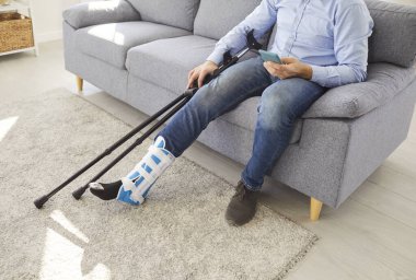 Engelli bir adam, gündelik giysiler içinde koltukta elinde cep telefonu tutan koltuk değnekleriyle oturuyor. Yaralı bacağı alçılı ya da bastonlu akıllı telefon kullanan adam..