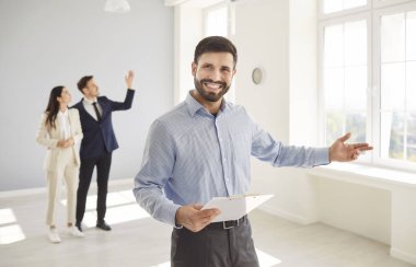Genç sakallı, gülümseyen bir emlakçının portresi mutlu evli bir çiftle yeni boş bir evde kameraya neşeli bir şekilde bakıyor. Yeni daireler gösteren profesyonel emlakçı..
