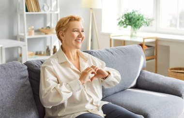 Yaşlı mutlu kadın evdeki bir kanepede oturuyor ve elleriyle kalp işareti yapıyor. Sevgi, şefkat, tutku ve sevgiyi yansıtan an an, olumlu duyguların yumuşaklığını vurguluyor..