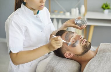 Kadın güzellik uzmanı, güzellik salonunda yakışıklı erkeklerin yüzüne sürtünerek kozmetik kil maske takıyor. Kozmetik uzmanı erkek müşteriye güzellik prosedürü uyguluyor. Sağlık ve cilt bakımı kavramı.