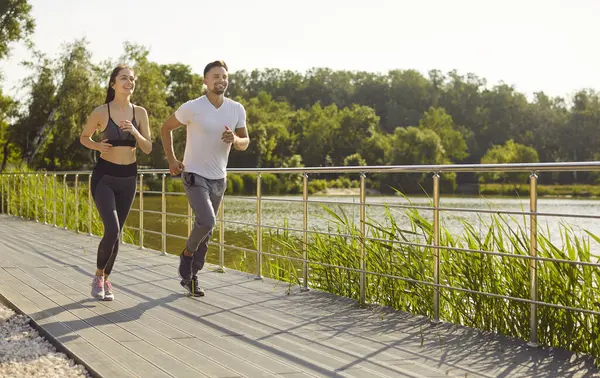 Göl kenarında güneşli bir gün, erkek ve kadın bir yolda birlikte koşuyor, spor yapıyor, spor yapıyor, sağlıklı yaşam tarzını sürdürüyor, sabah sporu planlarını gerçekleştiriyor.