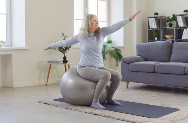 Kıdemli yaşlı kadın dengeli topun üzerinde oturuyor, yoga minder fitness ve sağlıklı gün oturma odasında egzersiz yapıyor ve esniyor, yaşlı kadın rehabilitasyon, fiziksel fonksiyon, hareketlilik için egzersiz yapıyor. 