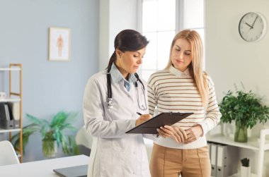 Laboratuvar önlüklü kadın doktor elinde bir dosya, hastaya tıbbi detayları anlatıyor. Muayene sırasında genç bayana danışmanlık yapan profesyonel doktor ya da kardiyolog.