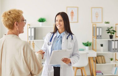 Beyaz laboratuvar önlüklü kadın doktor. Parlak ofisteki yaşlı kadın hastayla gülümseyip el sıkışıyor. Neşeli kardiyolog elinde bir not panosuyla danışma sırasında dostça sohbete girişiyor..