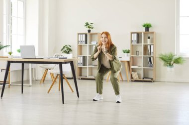 Genç gülümseyen bir kadının tam boy portresi. Ofiste ayakta oturma egzersizleri yapıyor. Neşeli bayan şirket çalışanı bir işe ara verdiğinde sağlık için egzersiz yapıyor..