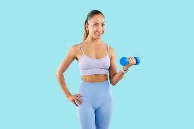 Spor giyim sektöründe gülümseyen güçlü ve mutlu bir kadının portresi yükselmiş bir halterle duruyor. Atletik vücutlu spor eğitmeni mavi arka planda poz veriyor. Spor sporu konsepti.