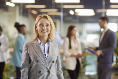 Toplantı odasında gülümseyen sarışın profesyonel şirket çalışanının portresi. Kurumsal eğitimden ya da toplantıdan sonra ayakta duran ve arka planda ekiple kameraya bakan kendinden emin bir kadın..