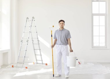 Boyalı silindirli ve elinde kovayla beyaz arka plandaki kameraya bakan kendine güvenen genç profesyonel ressamın tam boy portresi. Müteahhit tadilat yapıyor. Onarım kavramı.