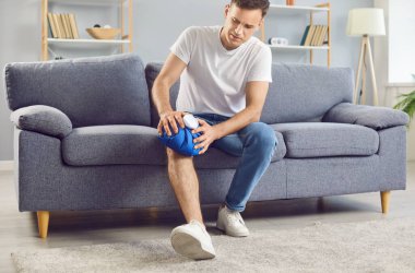 Evinde bacağını yaralayan talihsiz bir adam ağrıyı hissediyor ve yaralı dizine buz torbası tutuyor. Yüzünde rahatsızlık ifadesi olan bir adam kanepede oturuyor ve ağrı kesici olarak serin jel paketi uyguluyor..