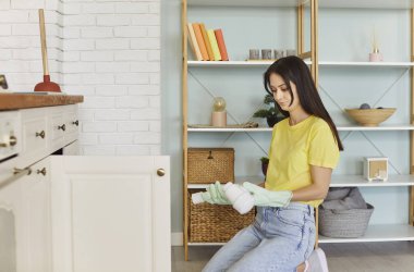 Lastik eldivenli bir kadın mutfakta yerde diz çöker, elinde endişeli ya da hayal kırıklığına uğramış bir ifadeyle kırık bir boru tutar. Tesisat sorunu, beklenmedik ev işi acil durum, profesyonel tamir hizmetlerine ihtiyaç var.