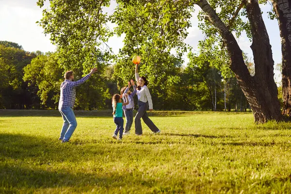 Mutlu gülümseyen dört kişilik aile, yaz tatillerinde yeşil çimenlerin üzerinde parkta top oynuyorlar. Gündelik giyinmiş çocuklu genç ebeveynler doğada eğleniyorlar..