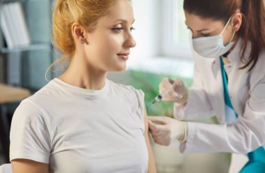 Kadın hasta aşı oluyor ya da muayenehanedeki doktordan aşı oluyor. Maskeli ve eldivenli sağlık uzmanı kliniğe rutin bir tıbbi ziyaret sırasında genç bayana iğne yapıyor..