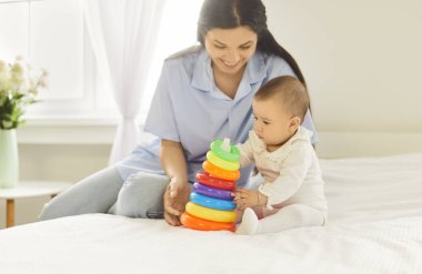 Mutlu gülümseyen anne yatakta oturup şirin bebeğinin renkli halka piramit oyuncağıyla oynamasını izliyor. Sevimli bebek öğrenme ve evdeki eğlenceli aktivitelerle iyi motor becerileri geliştirme.