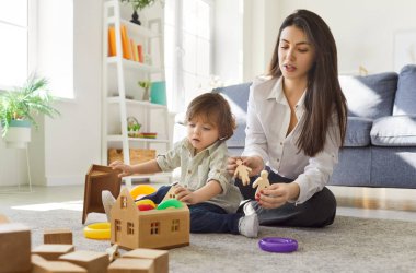 Ebeveynlik ve çocuk yetiştirme kavramı. Önemseyen bir anne küçük oğluyla oynar, ahşap bir ev yapar, rahat bir odada sıcak zeminde poz verir. Anne ve çocuk evde birlikte oynuyorlar..