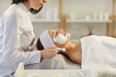 Profesyonel kozmetolojistin, kaplıca salonundaki kanepede uzanan genç bir kadına nemlendirici maske takması. Kadın müşteri güzellik prosedürünü uyguluyor. Sağlık, cilt bakımı ve yüz bakımı.