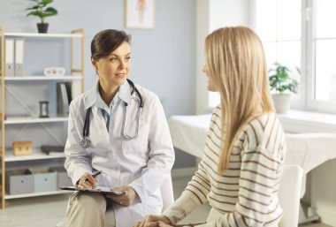 Laboratuvar önlüklü kadın doktor, tıp ofisinde oturuyor, dikkatle genç bayanı dinliyor ve notlar alıyor. Bakım hekimi hastaya profesyonel danışmanlık yapıyor, sağlık sorunlarını tartışıyor..