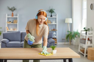 Ev temizliği, mutlu kızıl saçlı bir kız, kulaklık takan bir kadın, ahşap masa yüzeyini yıkayan eldivenler, temizlikçi, günlük ev işleri yapan bir ev kadını, toprağı temizleyen, temizleyen bir kadın.