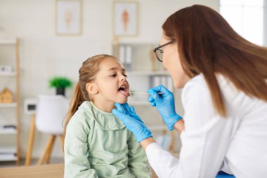 Kadın gülümseyen çocuk doktoru sağlık kliniğinde küçük kız hastaların boğazını muayene ediyor. Doktor kadın otolarolog muayenehanedeki hasta bir çocuğun ağız boşluğunu muayene ediyor..