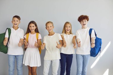 Mutlu ilkokul çocukları baş parmağını kaldırır. Sırt çantaları gri stüdyo duvarında duran ve baş parmaklarını yukarı kaldıran günlük giysili, neşeli erkek ve kadın çocukların toplu portresi.