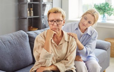 Endişeli yetişkin kızı, üzgün yaşlı annesini rahat bir evde, kanepede otururken rahatlatıyor. Anne ve kızının duygusal desteği. Bu an empati, bakım ve jenerasyon bağlantısını gösteriyor.. 