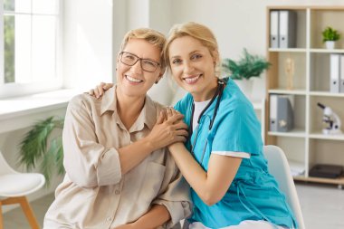Tıbbi bir ziyaret sırasında yaşlı bir kadınla el ele tutuşan gülümseyen hemşire, modern, parlak bir sağlık ofisinde bakım ve destek gösteriyor. Arkadaş canlısı bakıcı ve yaşlı bir hasta..
