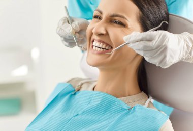 Modern dişçilik kliniğinde tıbbi muayene sırasında ağzı açık olan kadın hastaların yakın plan portresi. Dişçi, dişçi aletleriyle genç kadın dişlerini inceliyor. Diş sağlığı hizmetleri.