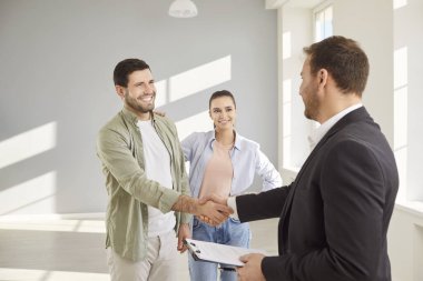 Emlakçıyla el sıkışan çift, emlak ya da daire alma ya da kiralama anlaşmasını onaylıyor. Takım elbiseli bir emlakçı, pano tutuyor ve mutlu beyaz bir adamla el sıkışıyor. Kadının yanında duruyor..