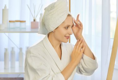 Beyaz bornoz ve havlu giymiş güzel bir kadının portresi banyodaki aynaya bakarak sabah güzellik ameliyatı yapıyor. Vücut sağlığı ve cilt bakımı kavramı.