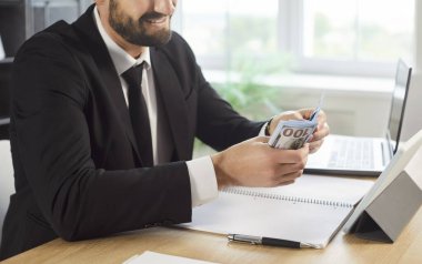 Yüzlerce dolarlık banknotları elinde tutan gülümseyen işadamının makamda paraları sayarken çekilmiş görüntüsü. Takım elbiseli bir erkek girişimci dizüstü bilgisayarla masada oturuyor, gelir hesaplıyor veya bütçeyi yönetiyor..