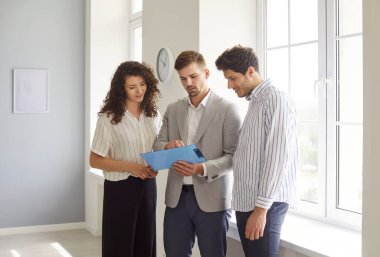 Genç, mutlu, gülümseyen bir çift erkek ve kadın profesyonel emlakçı ile modern boş dairelerde duran sözleşmeyi tartışıyor. Gayrimenkul ve emlak kavramı.