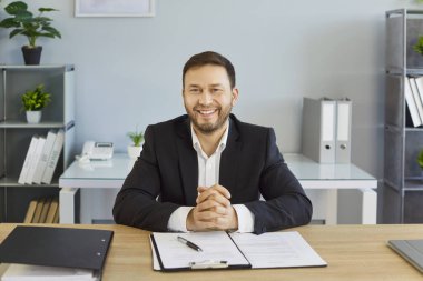 Resmi takım elbiseli yakışıklı, güler yüzlü iş adamının portresi. Çalışma yerinde evrakları var ve kameraya bakıyor. Ofiste çalışan mutlu erkek şirket çalışanı.